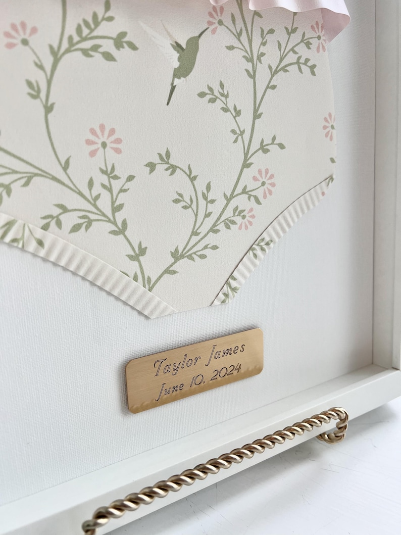 May include: A white frame with a gold trim and a gold plaque that reads "Taylor James June 10, 2024". The frame contains a white background with a floral pattern and a small hummingbird.