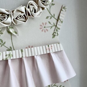 May include: A pink and white paper dress with a ruffled skirt and a floral pattern. The dress is decorated with a row of white paper roses.