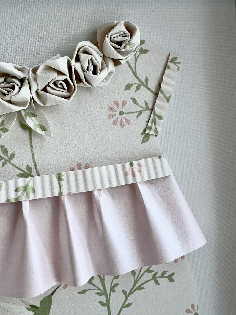 May include: A pink and white paper dress with a ruffled skirt and a floral pattern. The dress is decorated with a row of white paper roses.