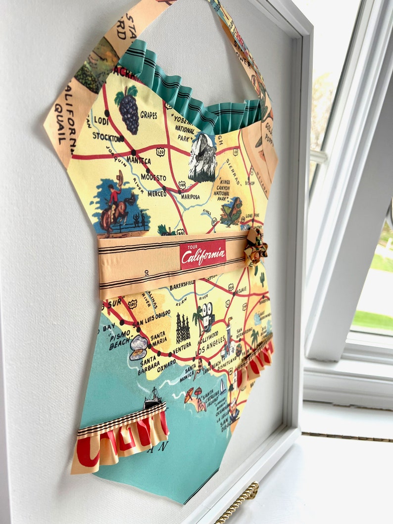 Pu&ograve; includere: Opera d'arte incorniciata a forma di costume da bagno, con una mappa vintage della California. La mappa &egrave; gialla, verde e blu, con linee rosse. Rifiniture a balze e un cinturino.