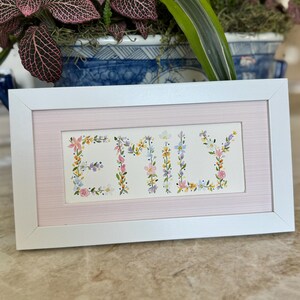 May include: A framed artwork featuring the name "EMILY" spelled out in colorful floral letters. The artwork is set against a pink background and framed in a white rectangular frame. The piece is displayed on a light-colored surface.