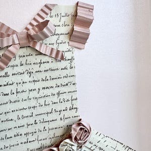 May include: A vintage letter with handwritten text in French, partially covered by a pink ribbon and paper roses. The text reads "le 15 Juilles fra rom clore s thegament Syndies of quite onper les bois r&eacute;cepaines &agrave; son hauffe nelle existait de deja mue instance mtre les i araitrait qu'an m&eacute;pris dela derni&egrave;re ordonn. Dans la force pour y exercer &theta;&alpha; &delta;&iota;ό&nu;&omicron; &delta;'&omicron;. rent dress&eacute;s sum le requisition des Officiers qui avaient coup&eacute; des bois dans la dite foret Fantua, le Conseil municipal dugrand Abay. anse on in aiss proue les d&eacute;lingnans faire de levas tithes appuy&eacute;s de la possession; que e des proc&eacute;s a erbans monregalement as Communes S'gos unbleness of renve"
