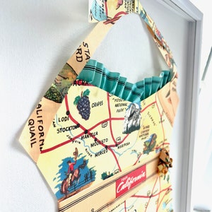 Pu&ograve; includere: Opera d'arte incorniciata con una mappa vintage della California. La mappa &egrave; stampata su tessuto e modellata a forma di abito. Il design include uno sfondo giallo chiaro con dettagli rossi e blu e il testo "TOUR California".