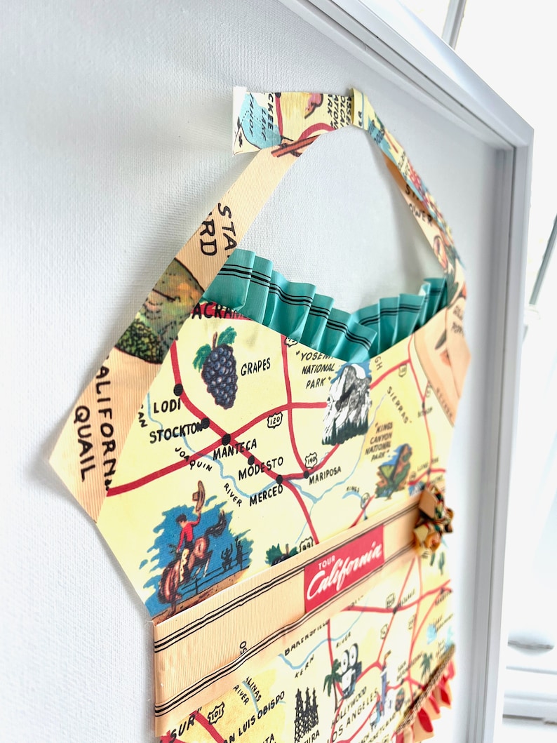 Pu&ograve; includere: Opera d'arte incorniciata con una mappa vintage della California. La mappa &egrave; stampata su tessuto e modellata a forma di abito. Il design include uno sfondo giallo chiaro con dettagli rossi e blu e il testo "TOUR California".
