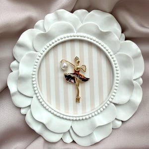 May include: A gold-toned ballerina brooch featuring a dark red skirt with clear crystals and a single white pearl accent. The brooch is mounted on a circular, white, petal-shaped frame with a beige and white vertically striped background.