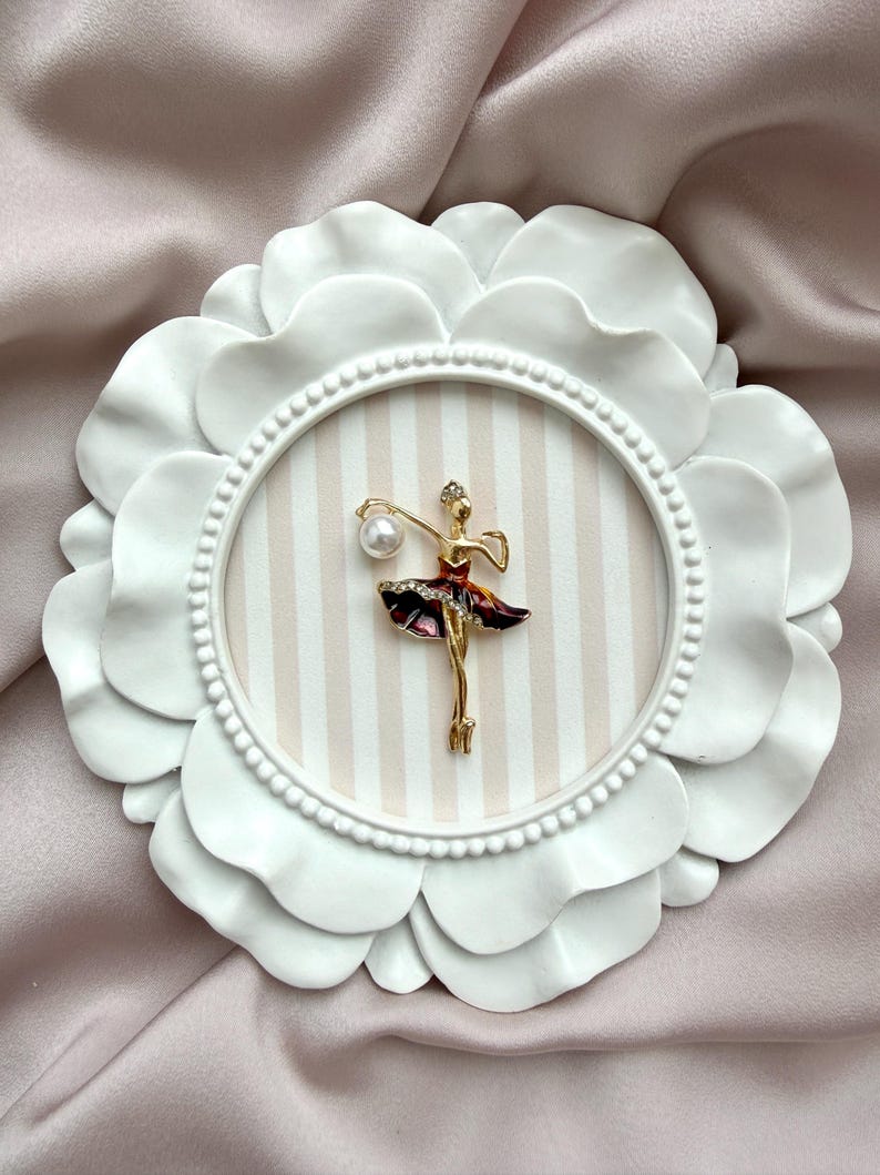 May include: A gold-toned ballerina brooch featuring a dark red skirt with clear crystals and a single white pearl accent. The brooch is mounted on a circular, white, petal-shaped frame with a beige and white vertically striped background.