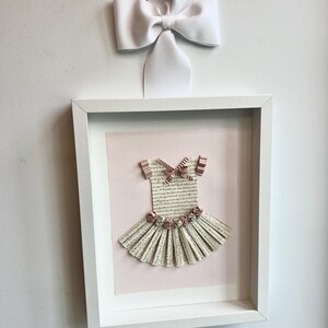 May include: A white frame with a pink background and a paper ballerina dress with pink flowers and a pink bow. The dress is made of paper and has a white background with black text.