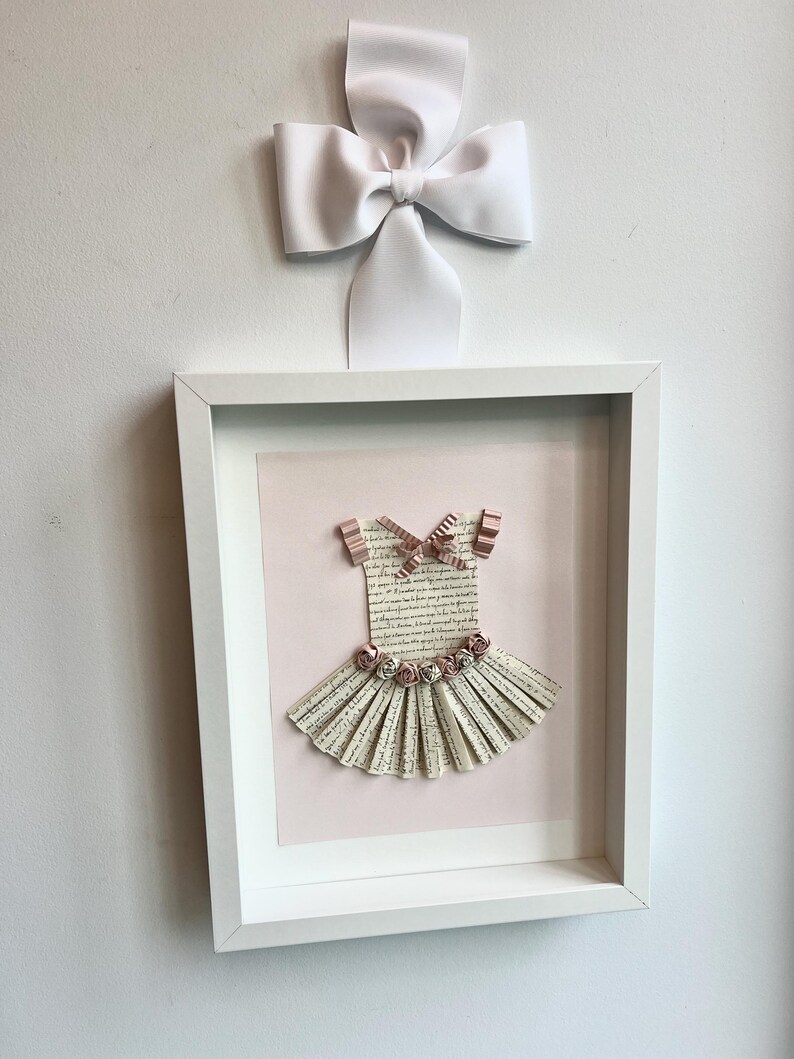 May include: A white frame with a pink background and a paper ballerina dress with pink flowers and a pink bow. The dress is made of paper and has a white background with black text.