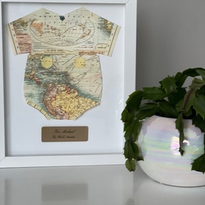 May include: A white framed wall art piece featuring a cutout in the shape of a baby bodysuit made from a vintage map of the world. Below the bodysuit is a rectangular plaque with the text For Michael The World Awaits printed in black ink.