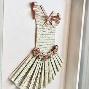 May include: A framed paper art piece depicting a ballerina's dress. The dress is made of folded paper with a pink ribbon and paper flowers.