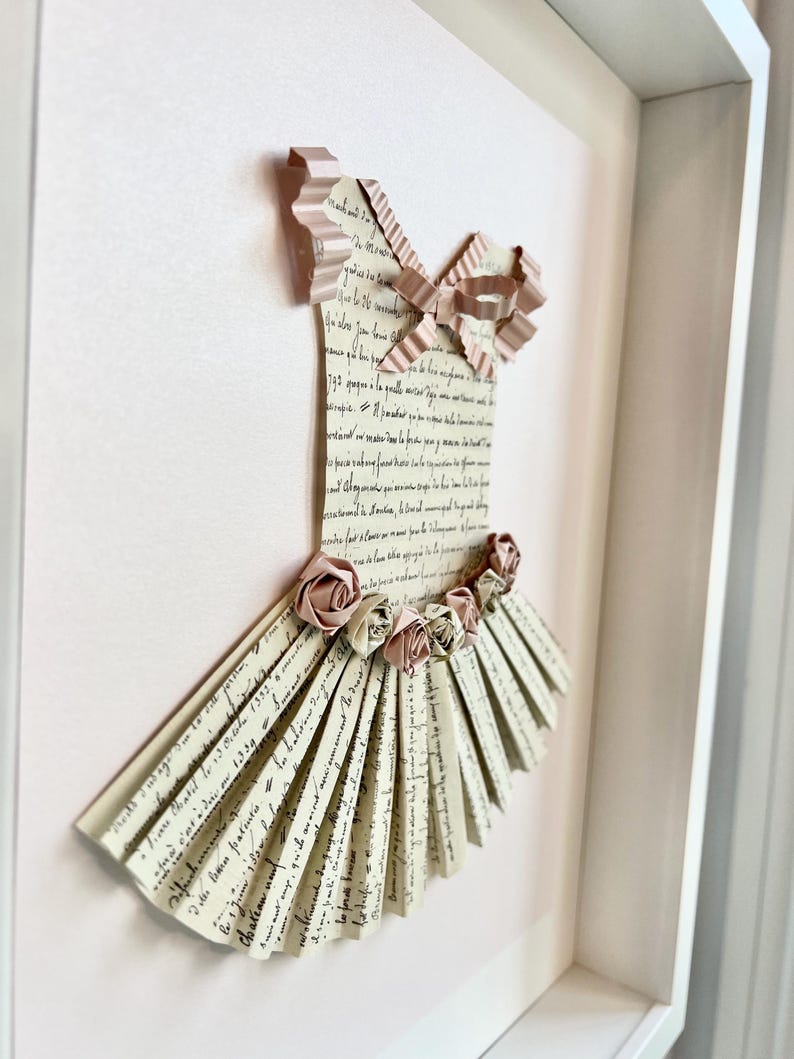 May include: A framed paper art piece depicting a ballerina's dress. The dress is made of folded paper with a pink ribbon and paper flowers.