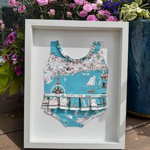 May include: A framed artwork featuring a teal and white swimsuit design. The swimsuit is decorated with a map of Cape Cod, sailboats, and a compass rose. The words "Cape Cod" are printed on the front. The artwork is displayed in a white frame.