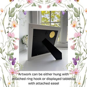 Pu&ograve; includere: Opera d'arte con cornice bianca con retro nero e cavalletto. Testo: L'opera d'arte pu&ograve; essere appesa con un gancio ad anello o esposta sul tavolo con un cavalletto. Bordo floreale con fiori rosa, viola e gialli.