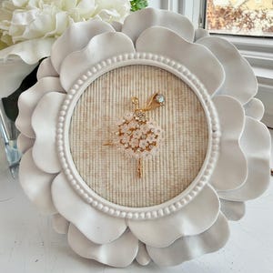 May include: A gold-toned ballerina brooch featuring a skirt made of small white floral accents and clear rhinestones, centered within a circular frame with a white petal-shaped border and a beige textured fabric backing.