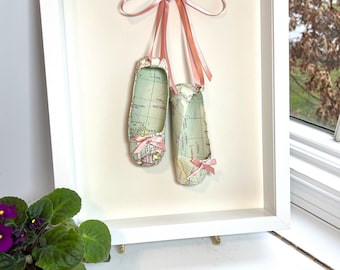 May include: A framed artwork featuring a pair of ballet shoes made from a vintage world map. The shoes are tied with a pink ribbon and are hanging from a nail in the frame.