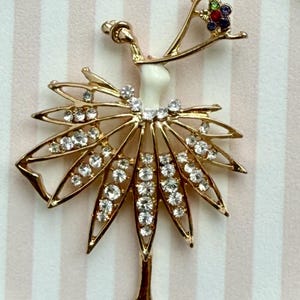 May include: A gold-toned metal brooch shaped like a dancer, featuring a white enamel torso, a skirt adorned with clear rhinestones, and a headpiece accented with small green, red, and blue stones. The dancer is posed with one arm raised.
