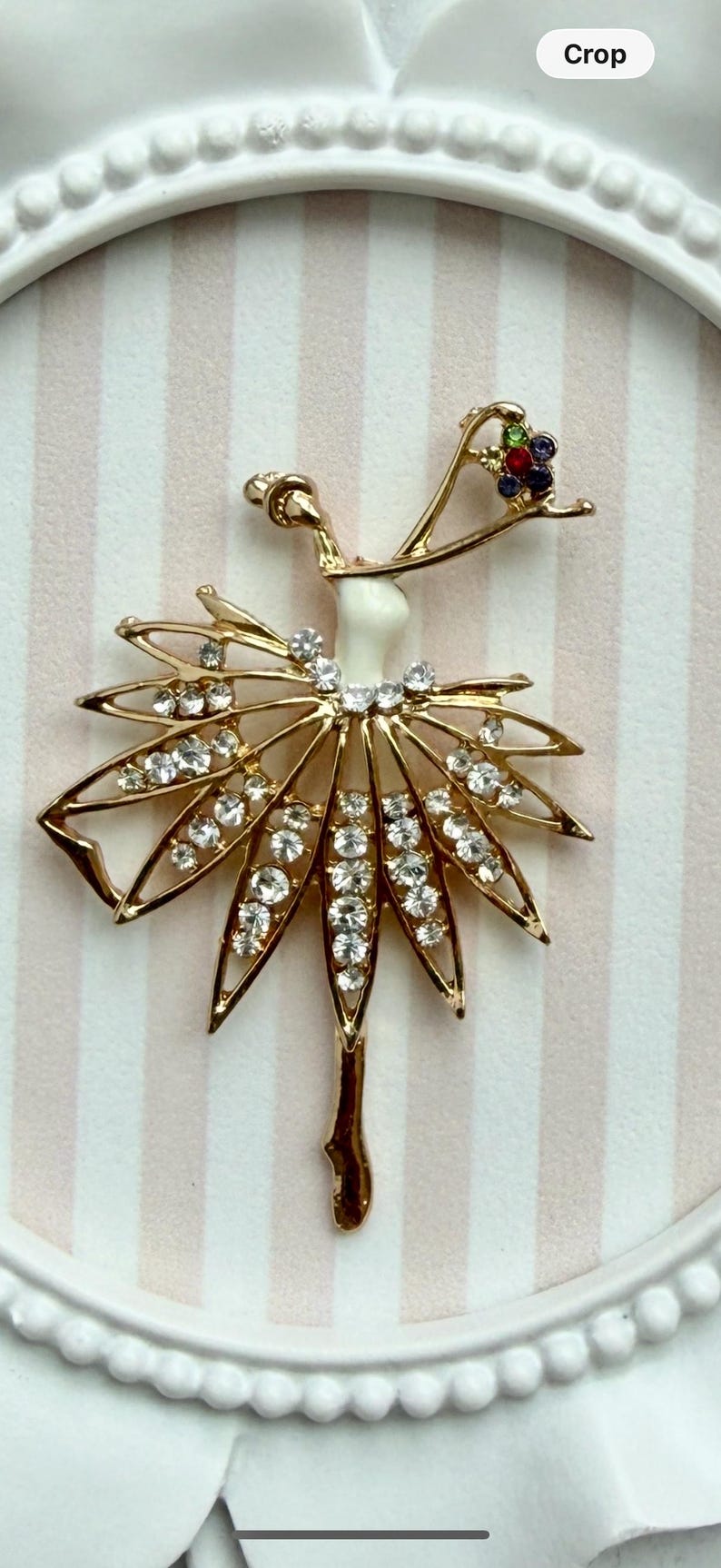 May include: A gold-toned metal brooch shaped like a dancer, featuring a white enamel torso, a skirt adorned with clear rhinestones, and a headpiece accented with small green, red, and blue stones. The dancer is posed with one arm raised.