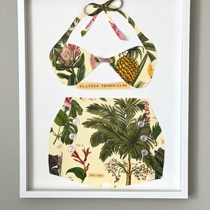 May include: A framed artwork featuring a white bikini made from vintage botanical illustrations of tropical plants. The bikini top has a bow at the center and the bottom has buttons. The text "Plantes Tropicales" is visible on the artwork.