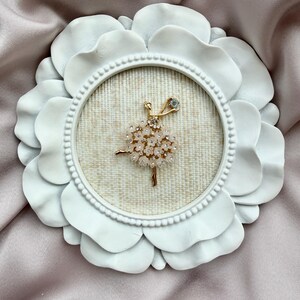 May include: A gold-toned brooch shaped like a ballerina, featuring a skirt made of small, pale pink flower-shaped embellishments and clear crystals. The brooch is displayed inside a round, white, petal-shaped frame with a textured beige fabric backing.