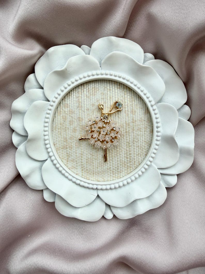 May include: A gold-toned brooch shaped like a ballerina, featuring a skirt made of small, pale pink flower-shaped embellishments and clear crystals. The brooch is displayed inside a round, white, petal-shaped frame with a textured beige fabric backing.
