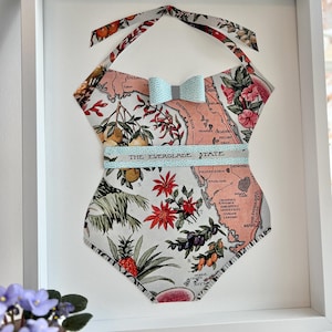 May include: Framed artwork featuring a fabric swimsuit design. The swimsuit is patterned with a map, fruits, and flowers, with the text "THE EVERGLADE STATE". A light blue bow and strap accent the design. The artwork is framed in a white frame.