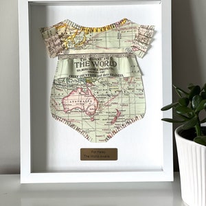May include: A white frame with a vintage map of the world cut out in the shape of a onesie. The map is in shades of brown, green, and blue. The text "The Century Atlas of The World" is visible on the map. A gold plaque at the bottom reads "For Haley The World Awaits."