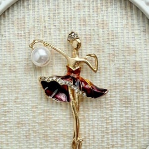 May include: A gold-toned ballerina brooch featuring a deep red enamel skirt accented with a row of clear rhinestones. The figure holds a single white faux pearl in one raised arm and wears a small rhinestone-encrusted headpiece.