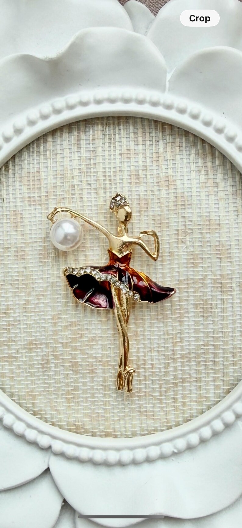 May include: A gold-toned ballerina brooch featuring a deep red enamel skirt accented with a row of clear rhinestones. The figure holds a single white faux pearl in one raised arm and wears a small rhinestone-encrusted headpiece.