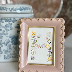 May include: A small, pink, scalloped-edge picture frame with a gold inner border. The frame holds a watercolor painting of the letter "H" decorated with colorful flowers and a small bee. A blue and white ceramic jar is in the background.