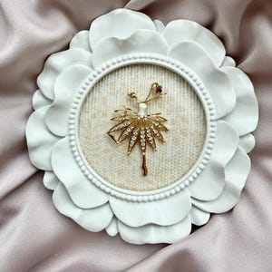 May include: A gold-toned ballerina brooch featuring clear rhinestones on the skirt and a small red stone accent, displayed inside a circular white frame with a scalloped petal-like border and a textured beige fabric backing.
