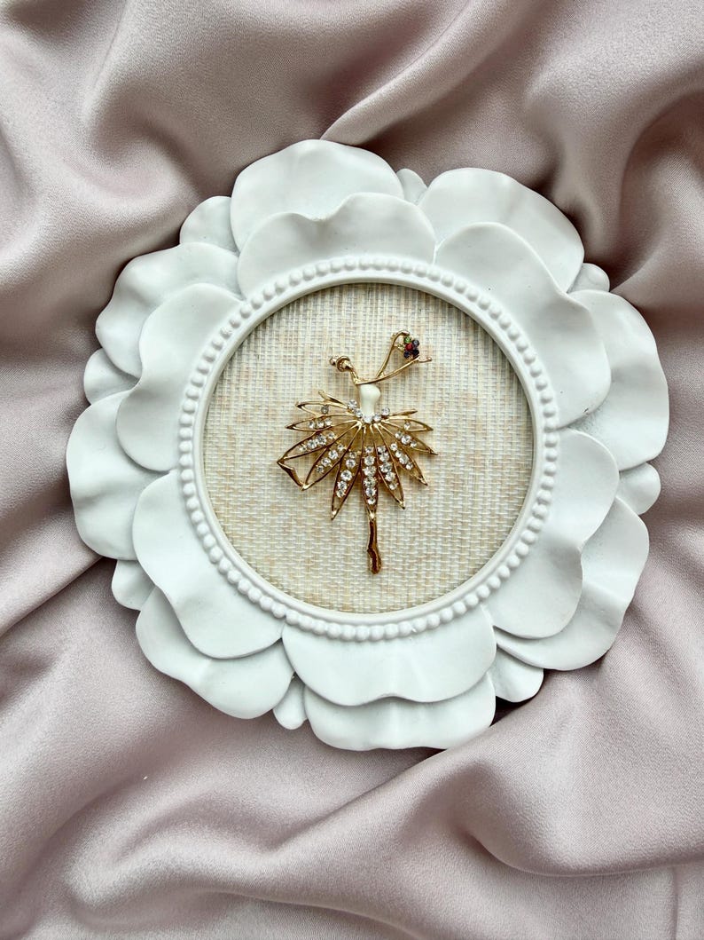 May include: A gold-toned ballerina brooch featuring clear rhinestones on the skirt and a small red stone accent, displayed inside a circular white frame with a scalloped petal-like border and a textured beige fabric backing.