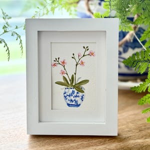 May include: A framed watercolor painting of a small pink flowering plant in a blue and white patterned pot. The painting is set in a white frame, with a wooden surface in the foreground and greenery in the background.