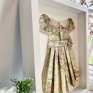 Puede incluir: Una obra de arte enmarcada que presenta un vestido de papel hecho de un mapa antiguo de Italia. El vestido est&aacute; decorado con un lazo rosa y un bot&oacute;n. El mapa muestra las regiones de Sicilia y Cerde&ntilde;a.