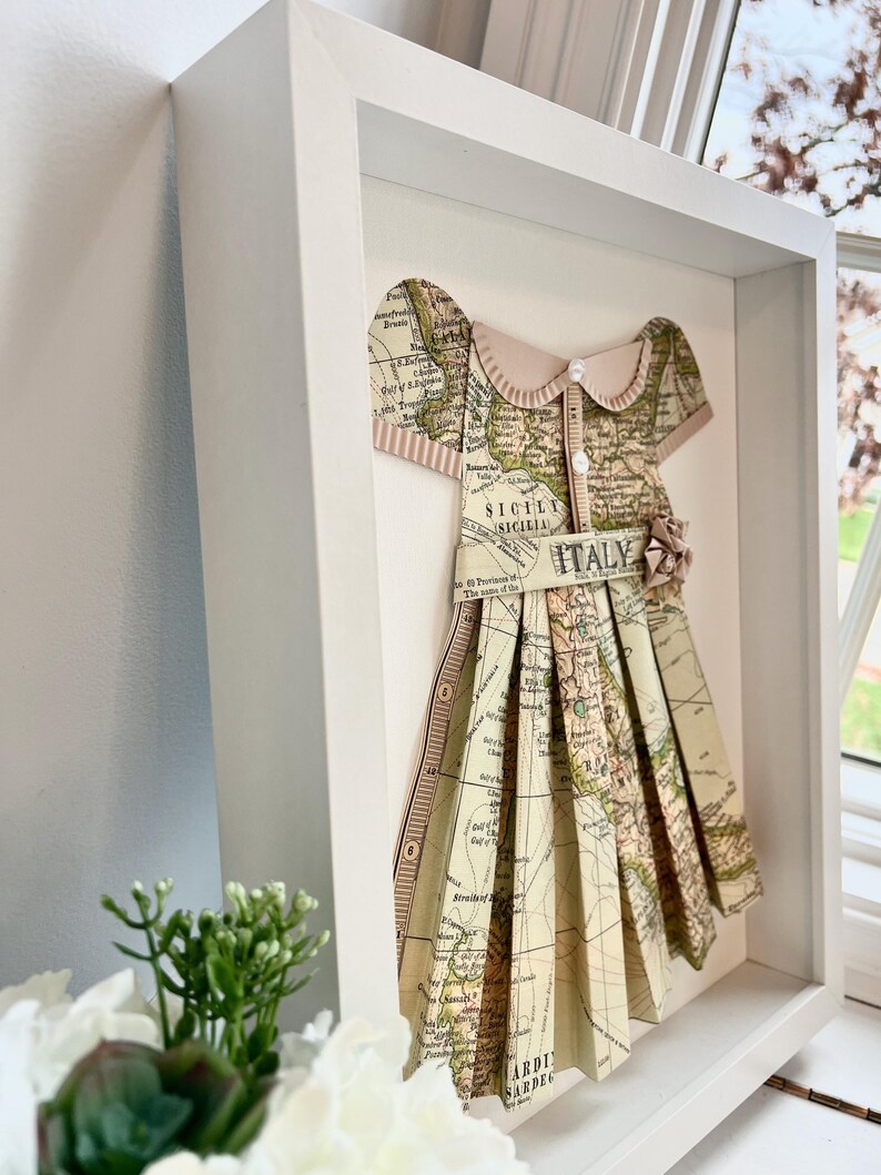Puede incluir: Una obra de arte enmarcada que presenta un vestido de papel hecho de un mapa antiguo de Italia. El vestido est&aacute; decorado con un lazo rosa y un bot&oacute;n. El mapa muestra las regiones de Sicilia y Cerde&ntilde;a.