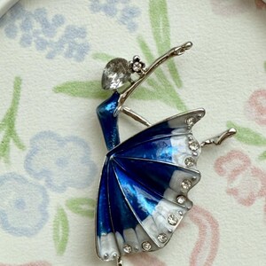 May include: A silver-toned brooch shaped like a ballerina in a pose. The figure features a blue and white enamel skirt accented with clear rhinestones along the hem and a single clear rhinestone forming the head of the dancer.