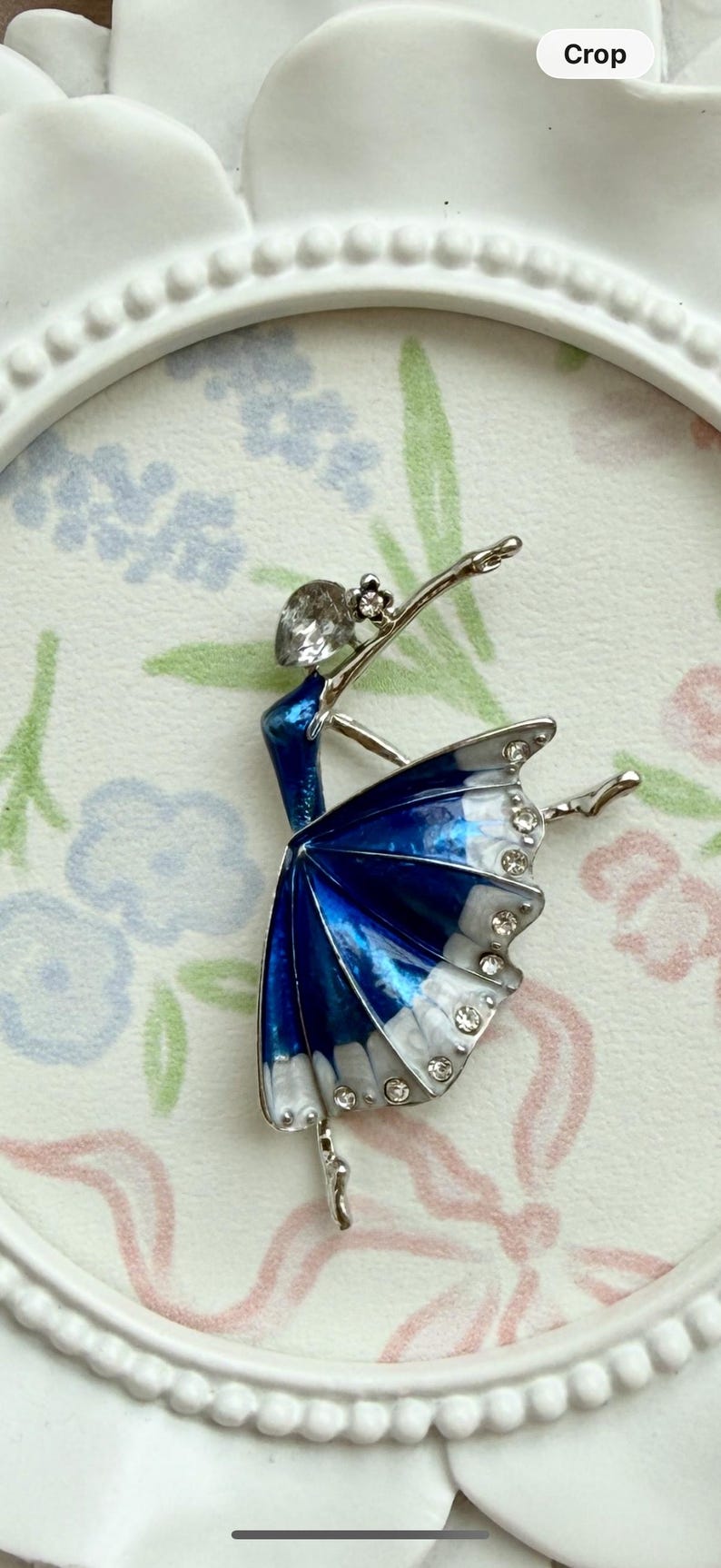 May include: A silver-toned brooch shaped like a ballerina in a pose. The figure features a blue and white enamel skirt accented with clear rhinestones along the hem and a single clear rhinestone forming the head of the dancer.