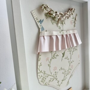 May include: A white frame with a paper cutout of a baby onesie with a floral pattern and pink ruffle. The onesie has a row of paper roses across the top. A gold plaque with the text "Taylor James June 10, 2023" is in the bottom right corner.