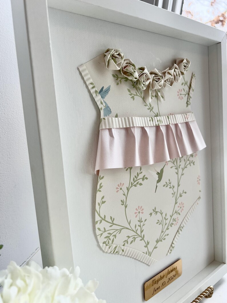 May include: A white frame with a paper cutout of a baby onesie with a floral pattern and pink ruffle. The onesie has a row of paper roses across the top. A gold plaque with the text "Taylor James June 10, 2023" is in the bottom right corner.