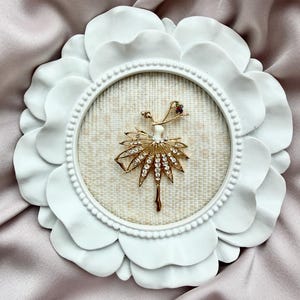 May include: A gold-toned brooch shaped like a dancer with a skirt made of clear rhinestones, set within a round white frame featuring a petal-like border and a textured beige fabric backing, displayed against a soft, draped fabric.