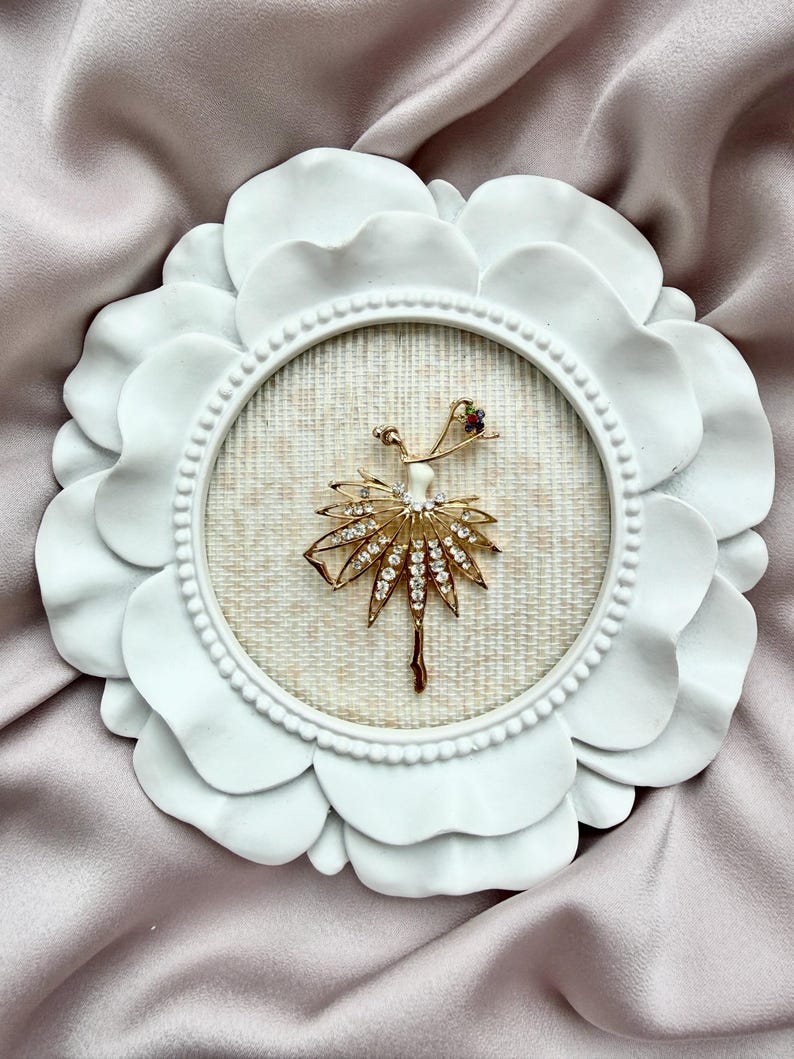 May include: A gold-toned brooch shaped like a dancer with a skirt made of clear rhinestones, set within a round white frame featuring a petal-like border and a textured beige fabric backing, displayed against a soft, draped fabric.