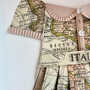 Puede incluir: Un vestido de estilo vintage hecho a partir de un mapa de Italia. El vestido tiene un corpi&ntilde;o marr&oacute;n con un cuello blanco y botones. La falda est&aacute; hecha de un mapa de Italia con las palabras "Italia" y "Sicilia" visibles. El vestido est&aacute; decorado con una cinta marr&oacute;n y una regla.