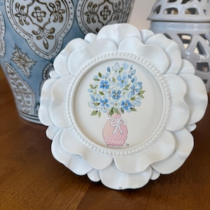 May include: A white, flower-shaped picture frame with a watercolor painting of blue flowers in a pink vase. The frame is set on a wooden surface, with a blue and white patterned vase in the background.