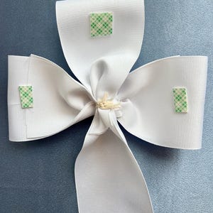 May include: A white satin bow with three green adhesive squares attached to the back.