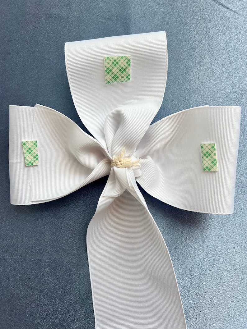 May include: A white satin bow with three green adhesive squares attached to the back.