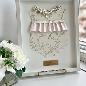May include: A white frame with a white background. Inside the frame is a paper cutout of a baby onesie with a pink ruffle and a floral pattern. The onesie has a row of white paper roses around the neckline. A gold plaque with the text "Taylor James June 10, 2022" is at the bottom of the frame.