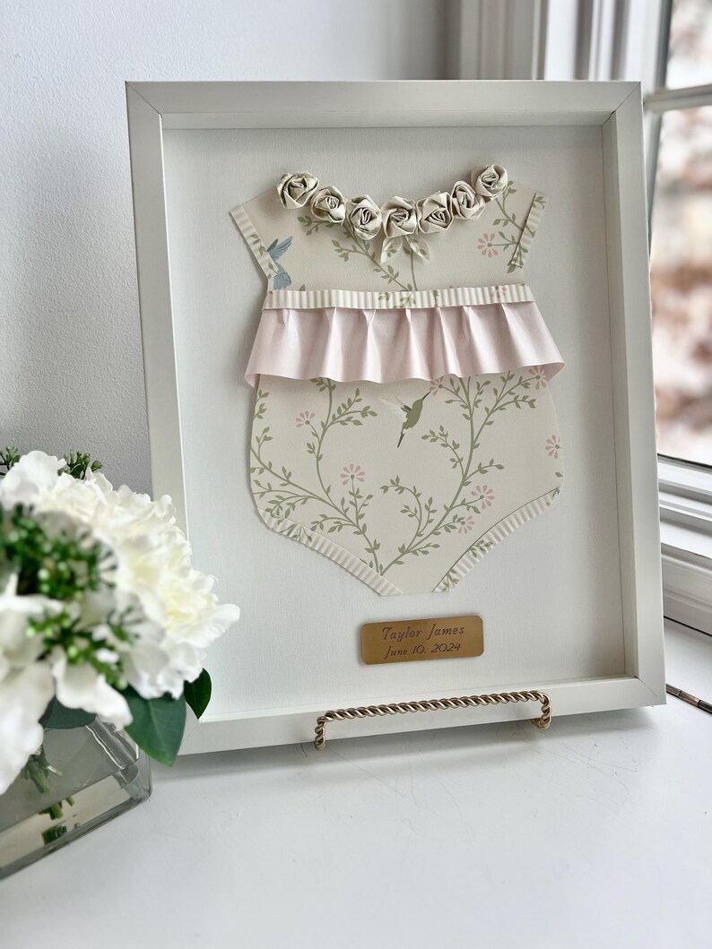May include: A white frame with a white background. Inside the frame is a paper cutout of a baby onesie with a pink ruffle and a floral pattern. The onesie has a row of white paper roses around the neckline. A gold plaque with the text "Taylor James June 10, 2022" is at the bottom of the frame.