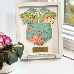 May include: A white picture frame with a world map cut out in the shape of a baby onesie. The map is a vintage style with blue and green colors. The frame has a gold plaque with the text "For Michael The World Awaits..."