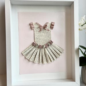 May include: A framed paper craft of a ballerina dress with a pink background. The dress is made of paper strips with a handwritten script on them. The dress has a pink bow and pink paper flowers.