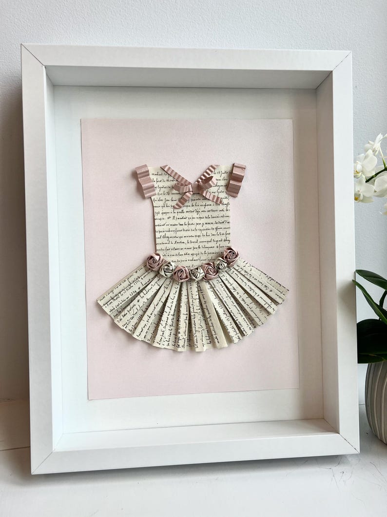 May include: A framed paper craft of a ballerina dress with a pink background. The dress is made of paper strips with a handwritten script on them. The dress has a pink bow and pink paper flowers.