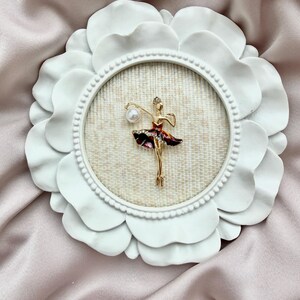 May include: A gold-toned ballerina brooch featuring a dark red skirt with clear crystals and a single white pearl accent, mounted inside a circular white frame with a scalloped edge and a textured beige fabric backing.